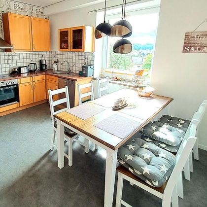 Eine helle und moderne Küche mit einem großen Esstisch und weißen Stühle. Der Raum ist gut beleuchtet und lädt zum Kochen und Essen ein.