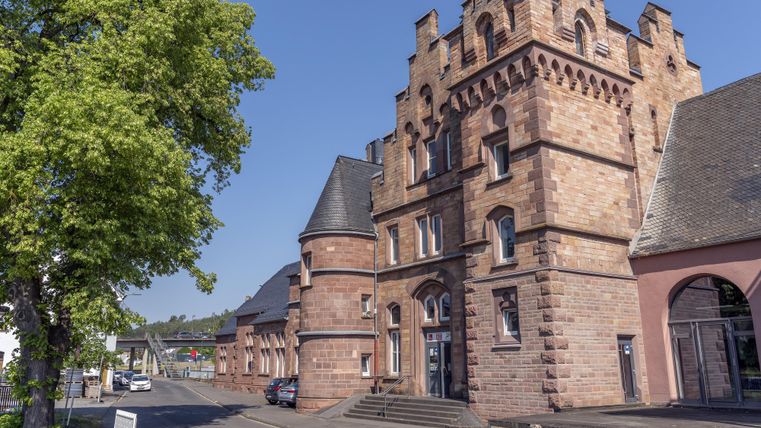 Historisches Bahnhofsgebäude in Gerolstein aus rotem Sandstein mit Türmen, daneben ein großer Baum und blauer Himmel.