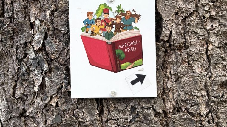 Ein bunter Aufkleber auf einem Baumstamm zeigt ein offenes Buch mit verschiedenen Figuren. Der Hinweis zeigt in Pfeilrichtung, vermutlich zu einem Pfad oder einer Attraktion.