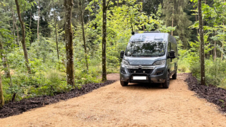 <p>Ein dunkler Wohnwagen fährt auf den Stellplatz auf. Der Stellplatz liegt mitten im Wald</p>