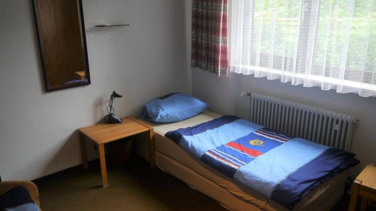Ein einfaches Zimmer mit einem Bett, einem Nachttisch und einem Spiegel. Durch das Fenster fällt natürliches Licht in den Raum.