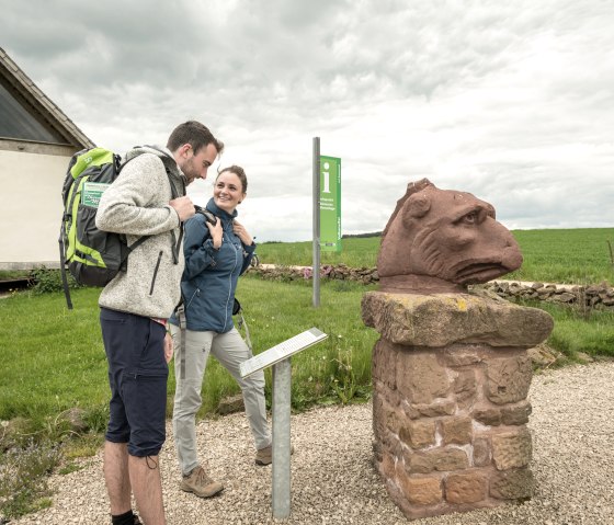 Zwei Wanderer stehen vor einer Steinskulptur auf einem Wanderweg in der Eifel. Im Hintergrund ist ein Gebäude und ein Informationsschild zu sehen., © Eifel Tourismus GmbH, Dominik Ketz