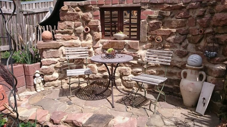 Un espace jardin confortable avec une table ronde et deux chaises en métal. Le mur est en pierre naturelle et il y a des éléments décoratifs tout autour.