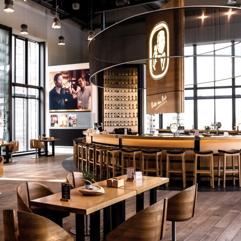 Restaurantraum der Bitburger Brauerei mit vielen Tischen, St&uuml;hlen und einer runden Theke in der Mitte. Gro&szlig;e Fensterw&auml;nde und passender Dekoration.