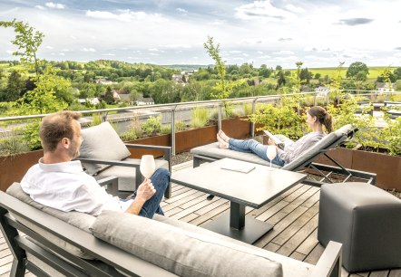 Een man en een vrouw zitten ontspannen op het modern ingerichte dakterras van een hotel, drinken wijn en kijken uit over het groene landschap.