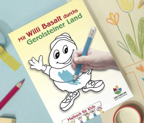 Cover page of the colouring book ‘With Willi Basalt through the Gerolstein region’ on a table full of colouring supplies and colourful sheets of paper.