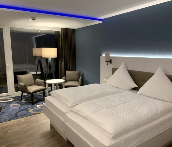 Modern eingerichtetes Hotelzimmer im Seehotel am Stausee mit einem gro&szlig;en wei&szlig; bezogenen Doppelbett, LED-Licht, Teppich und einem Beistelltisch mit St&uuml;hlen.