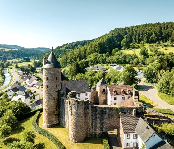 Luchtfoto van kasteel Bertrada in Mürlenbach, omgeven door groen landschap, een rivier en huizen. Het kasteel heeft een ronde toren en historische muren., © Eifel Tourismus GmbH, Dominik Ketz