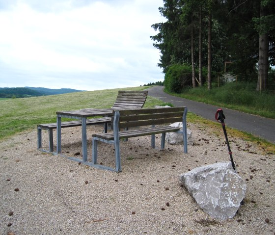 Picknickplatz mit Bänken und Tisch auf einem Hügel, daneben ein Wanderstock, im Hintergrund Wald und Wiese., © Touristik GmbH Gerolsteiner Land, Ute Klinkhammer