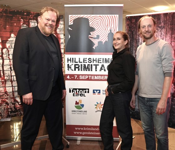 Zwei M&auml;nner und eine Frau stehen nebeneinader neben einem Banner zu den Hillesheimer Krimitagen und vor thematischen Leinw&auml;nden und l&auml;cheln in die Kamera.