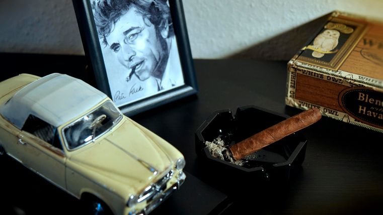 Une voiture miniature blanche se trouve à côté d'un cadre avec un dessin d'une personne en train de fumer. À côté, il y a un cendrier avec un cigare et une boîte de cigarettes.