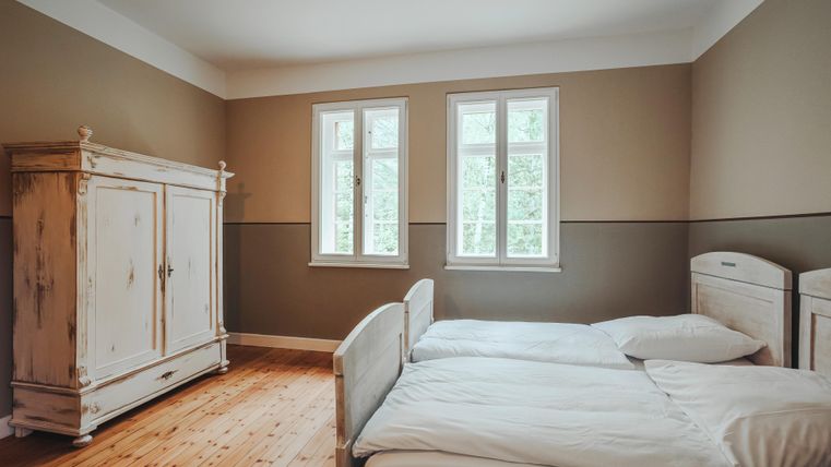 Dieses Bild zeigt ein Schlafzimmer mit zwei Einzelbetten und einem großen antiken Kleiderschrank. Durch zwei Fenster hat man einen Blick auf die Bäume draußen.