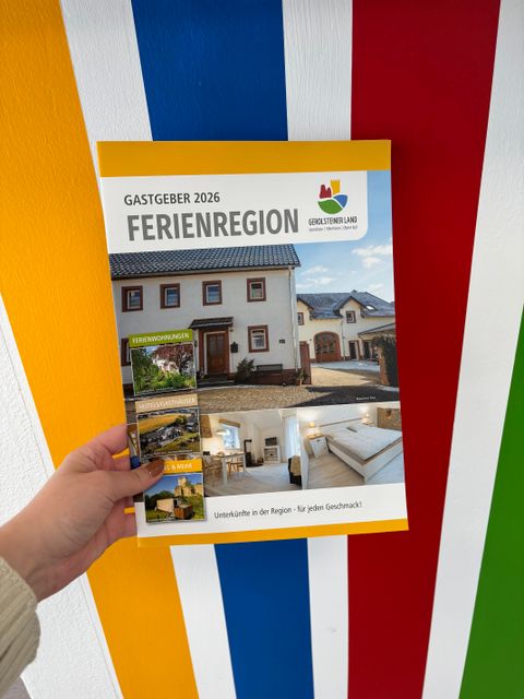 Eine Hand hält eine Broschüre über Unterkunftsbetriebe in der Ferienregion Gerolsteiner Land für 2026 vor eine Wand.