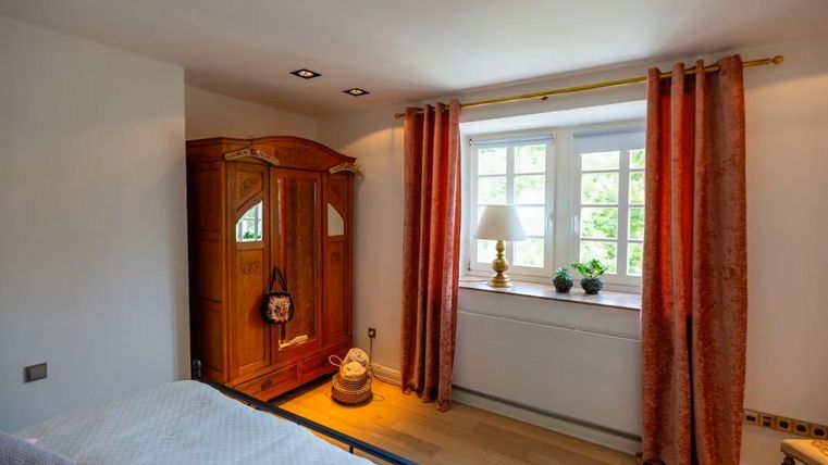 Ein helles Schlafzimmer mit einem großen Fenster und warmen Vorhängen. Ein Holzschrank und eine Nachttischlampe verleihen dem Raum eine gemütliche Atmosphäre.