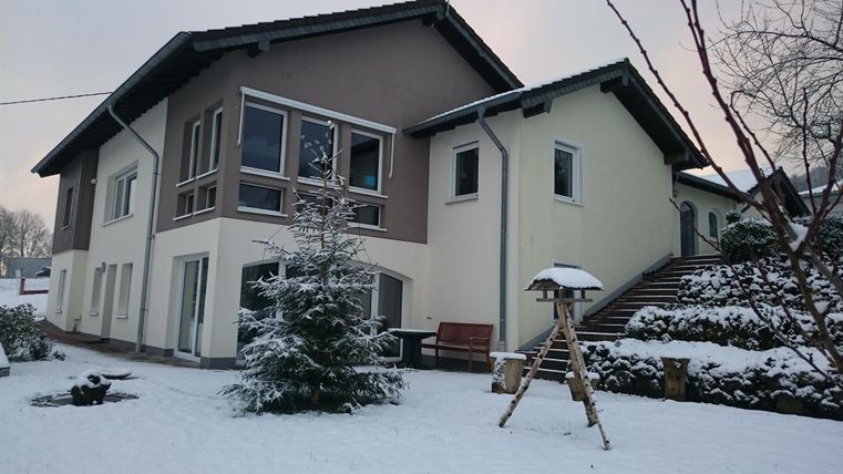 Ein modernes Haus im Schnee mit einer gepflegten Rasenfläche. Im Vordergrund steht ein kleiner Baum und eine Schaukel.