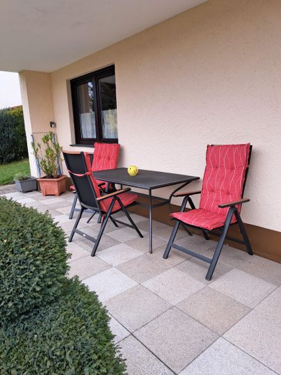 Eine Terrasse mit einem  Tisch und Gartenstühlen. Im Hintergrund sind grüne Pflanzen und ein gepflegter Rasen zu sehen.