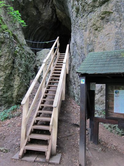 Wooden stairs lead to the dark entrance of the Buchenloch cave. To the right, there is an information board with a roof. The surrounding area is rocky and wooded.