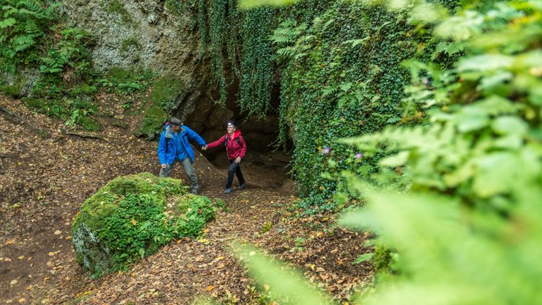 Zwei Wanderer kommen gemeinsam aus einer Höhle in einer massiven Felswand heraus in den angrenzenden Wald,