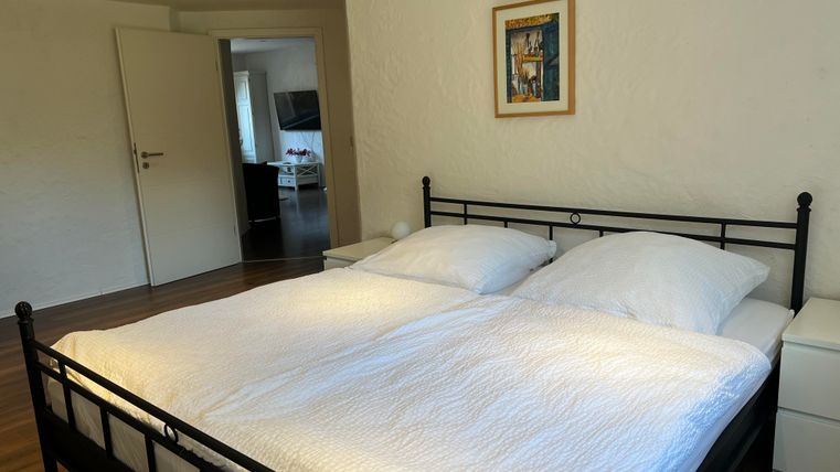 Een gezellige slaapkamer met een groot tweepersoonsbed en witte dekens. Op de achtergrond is een deur naar een andere kamer te zien.