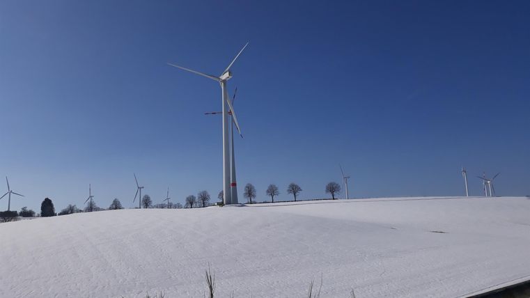 Eine Schneelandschaft mit Windkraftanlagen unter einem klaren blauen Himmel. Im Vordergrund sind vereiste Gräser zu sehen.
