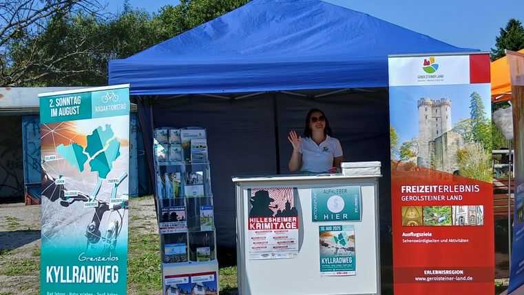 Informationsstand der Touristik GmbH Gerolsteiner Land am Radaktionstag "Grenzenlos Kyllradweg" mit Plakaten, Bannern und Flyern. Eine Mitarbeiterin winkt.