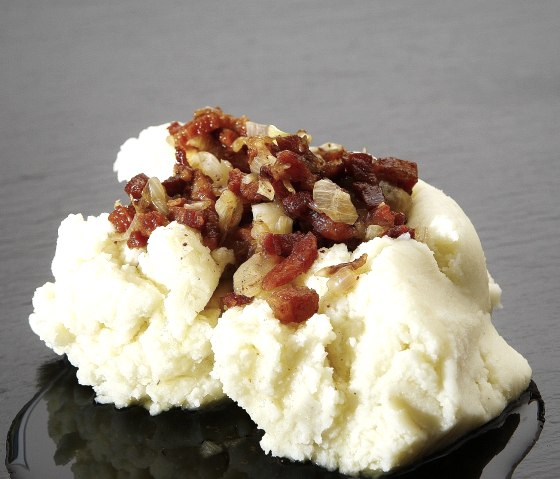 Ein Haufen Kartoffelpürree, auf dem Speck- und Zwiebelwürfel angerichtet sind.