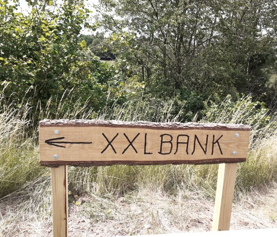 Holzschild mit Aufschrift 'XXL BANK' und Pfeil nach links, umgeben von Gras und Bäumen., © Touristik GmbH Gerolsteiner Land
