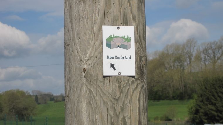 Holzpfosten mit Schild 'Maar Runde Auel' und Pfeil, im Hintergrund grüne Wiesen und Bäume.