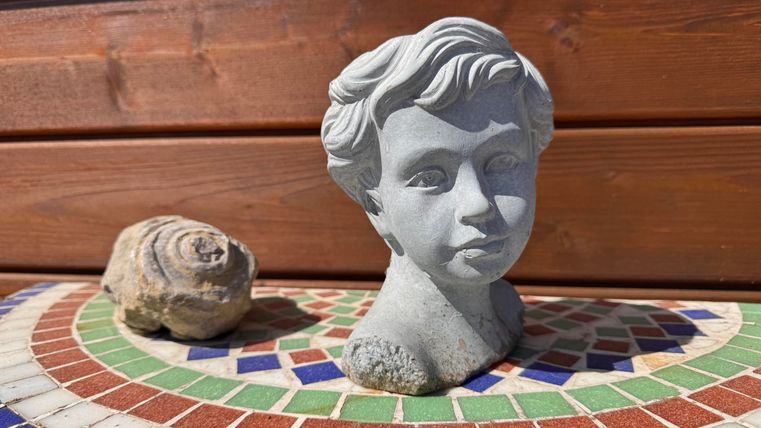 Eine Steinbüste eines Jungen steht auf einem bunten Mosaik-Tisch. Daneben liegt eine grobe Steinschnecke.