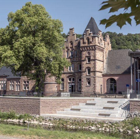 Außenfassade des Bahnhofs Gerolstein mit Treppe und Aufzug als Zugang zu den Gleisen hinter einem dicht bewachsenen Baum und neben dem Fluss Kyll.