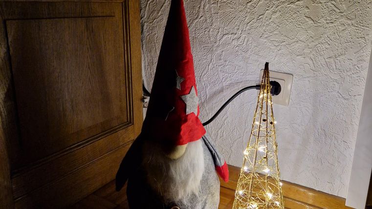 Een decoratieve kerstfiguur in de vorm van een gnoom staat naast een stralende, sprankelende kerstboom. De ruimte heeft een warme en feestelijke sfeer.