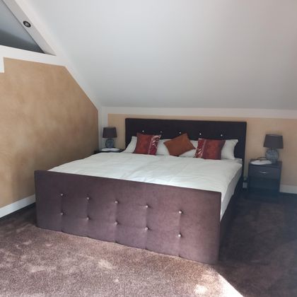 Ein gemütliches Schlafzimmer mit einem großen Doppelbett und dekorativen Kissen. Die Wände sind in warmen Farben gestrichen, und es gibt Nachttische mit Lampen.