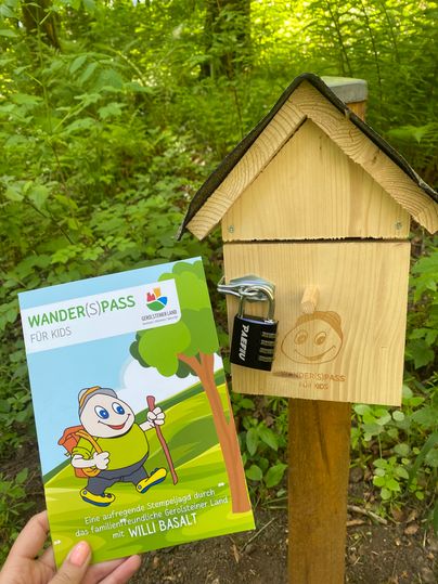 Das Prospekt "Wander(S)pass für Kids" wird hochgehalten vor einer dazugehörigen Stempelstation.