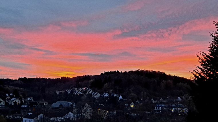 Ein malerischer Sonnenuntergang über einem ruhigen Ort, mit bunten Himmelstönen von Orange und Pink. Die Silhouetten der Häuser und Bäume verleihen der Szene eine friedliche Atmosphäre.