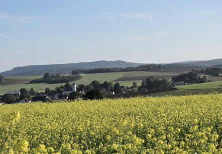 A vast rapeseed field stretches out in front of a village, surrounded by green hills and wind turbines under a clear sky., © Touristik GmbH Gerolsteiner Land