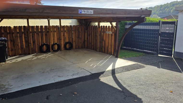Une aire de stationnement couverte avec des murs en bois et un portail d'entrée. À l'arrière-plan, des pneus et des conteneurs à déchets sont visibles.