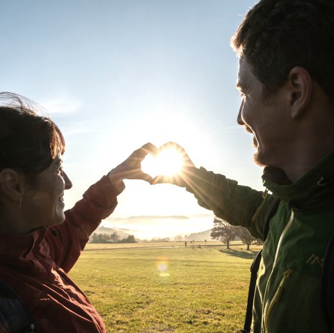 Two hikers on a vast meadow at sunrise form a heart shape with their hands.