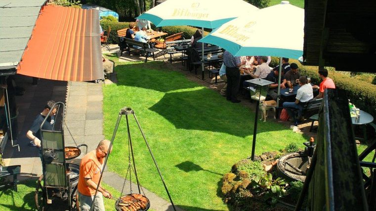 Ein Garten mit Grill, wo zwei Männer Fleisch zubereiten. Im Hintergrund sitzen Menschen unter Bitburger-Sonnenschirmen an Tischen.