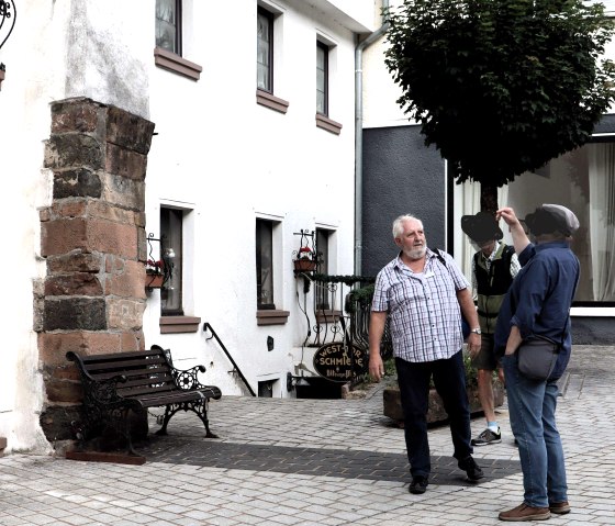 Twee mannen staan voor een oude boog. De een gebaart terwijl de ander luistert. Een boom en een gebouw op de achtergrond., &copy; Clara Zins-Groh&eacute;