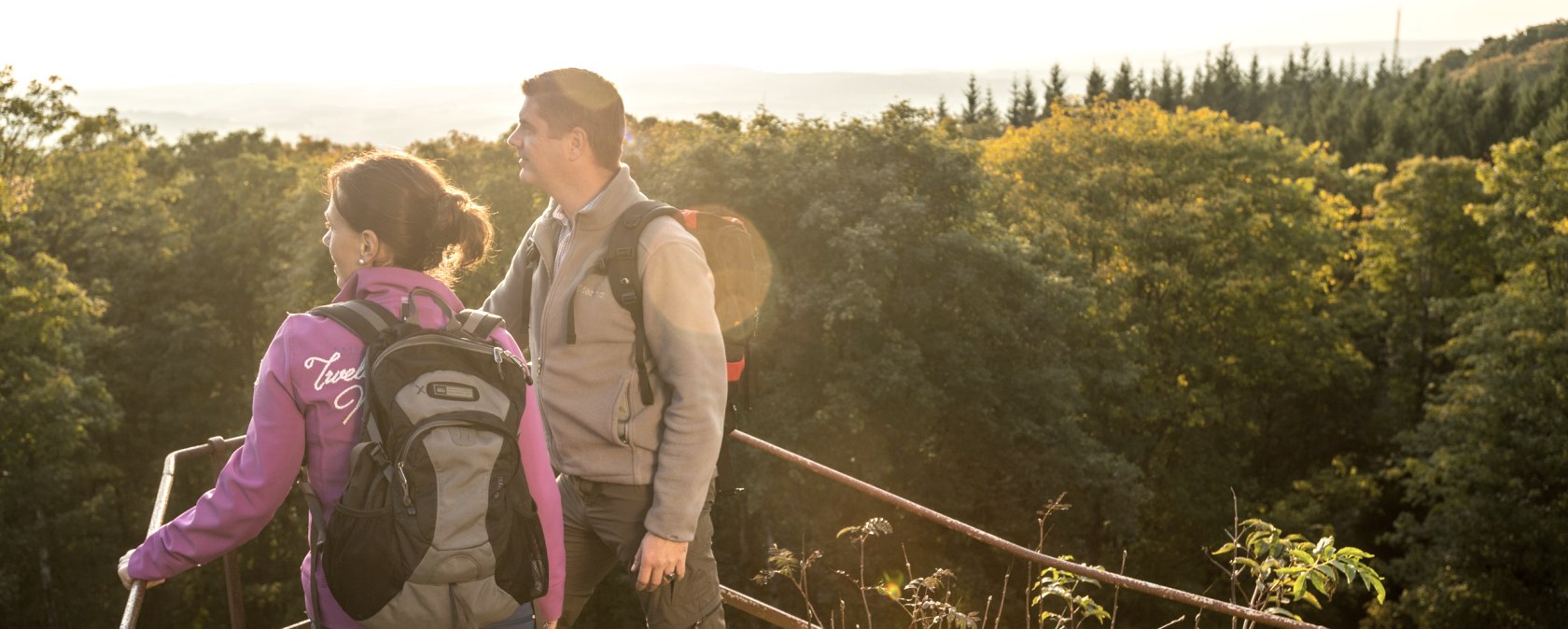 Zwei Personen stehen auf einem Felsen mit Blick auf einen Wald und H&uuml;gel im Hintergrund.
