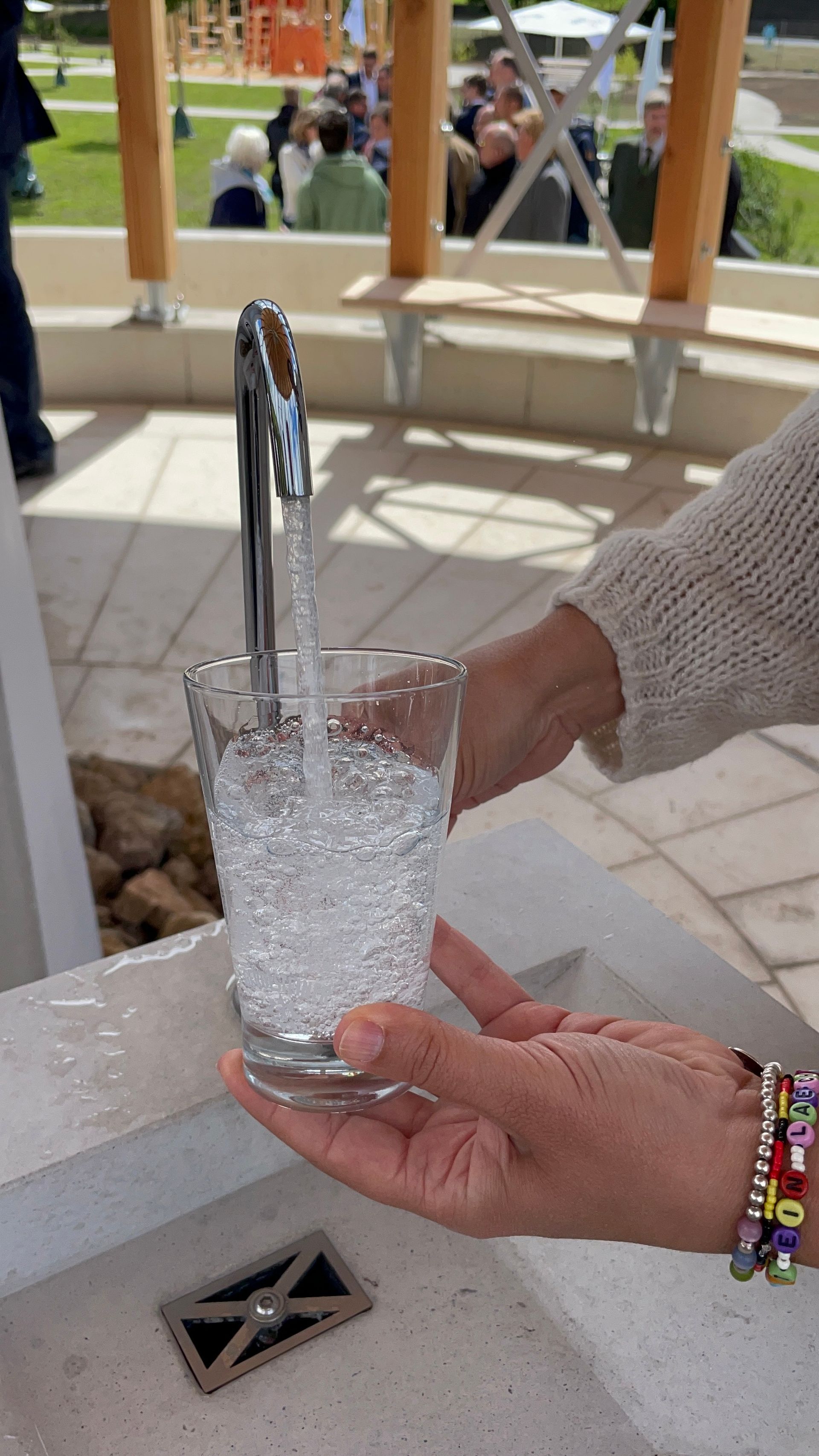 Ein Glas wird mit sprudelndem Wasser aus einem Wasserhahn gefüllt, im Hintergrund sind Menschen und ein Pavillon zu sehen.