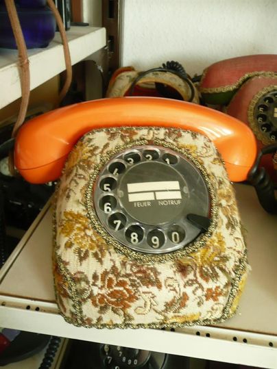 Ein orangefarbenes Wählscheibentelefon mit floralem Stoffüberzug auf einem Regal.