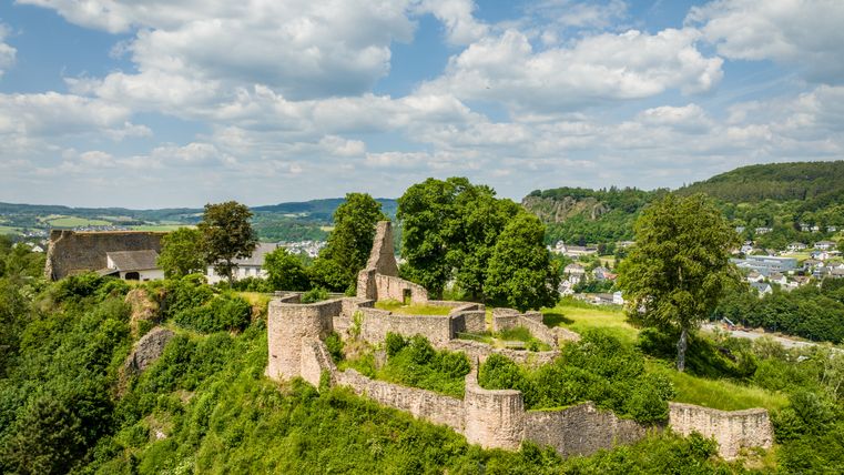 Luftaufnahme der Löwenburg in Gerolstein, umgeben von grüner Landschaft und Bäumen. Im Hintergrund ist die Stadt Gerolstein zu sehen, unter einem blauen Himmel mit Wolken.