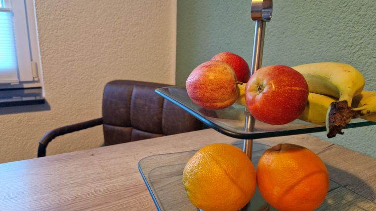 Ein schöner Obstständer mit Äpfeln, Bananen und Orangen steht auf einem Holztisch. Im Hintergrund ist ein Stuhl und eine Wand zu sehen.