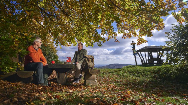 Deux personnes sont assises sous un arbre et profitent de la vue. Autour d'elles, on peut voir de feuilles d'automne colorées et un paysage paisible.