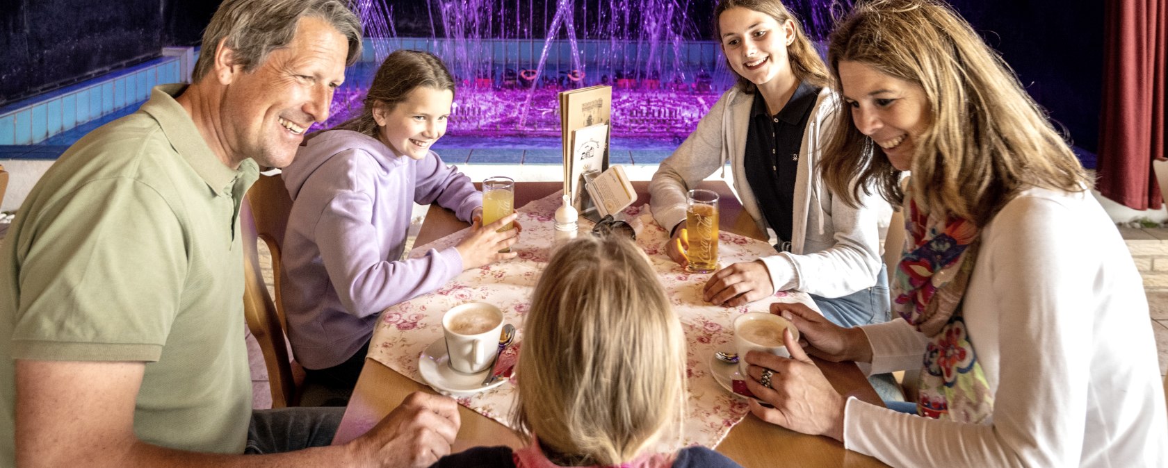 Een man en een vrouw zitten met hun drie dochters aan tafel en drinken koffie of frisdrank. Achter de familie spuit een kleurrijk waterorgel water in de lucht.