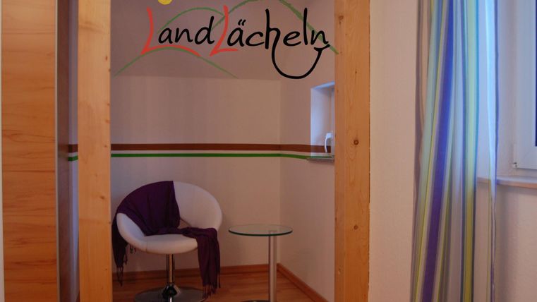 Ein gemütlicher Raum mit einem weißen Stuhl, einem Beistelltisch und bunten Vorhängen. An der Wand ist das Wort „Landlächeln“ kreativ gestaltet.