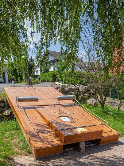 Große Holzkonstruktion in Form einer Mausefalle im Freien, umgeben von Bäumen und Häusern.