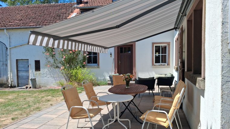 Ein gemütlicher Außenbereich mit einem Tisch und Stühlen unter einem Markisenbeschattung. Im Hintergrund ist ein traditionelles Haus mit weißen Wänden und grünen Pflanzen zu sehen.