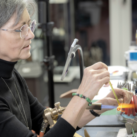 Eine Dame sitzt an einem Tisch und arbeitet konzentriert mit einem Brenner und Glas.